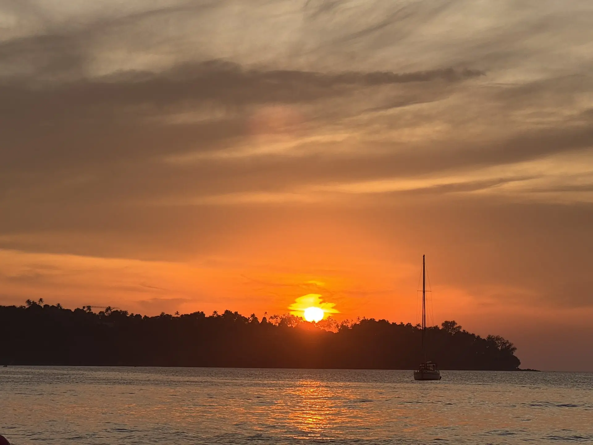Scenic sunset on Bang Tao Beach: an orange sun sets behind the silhouette of a hill, with a sailing yacht on the water