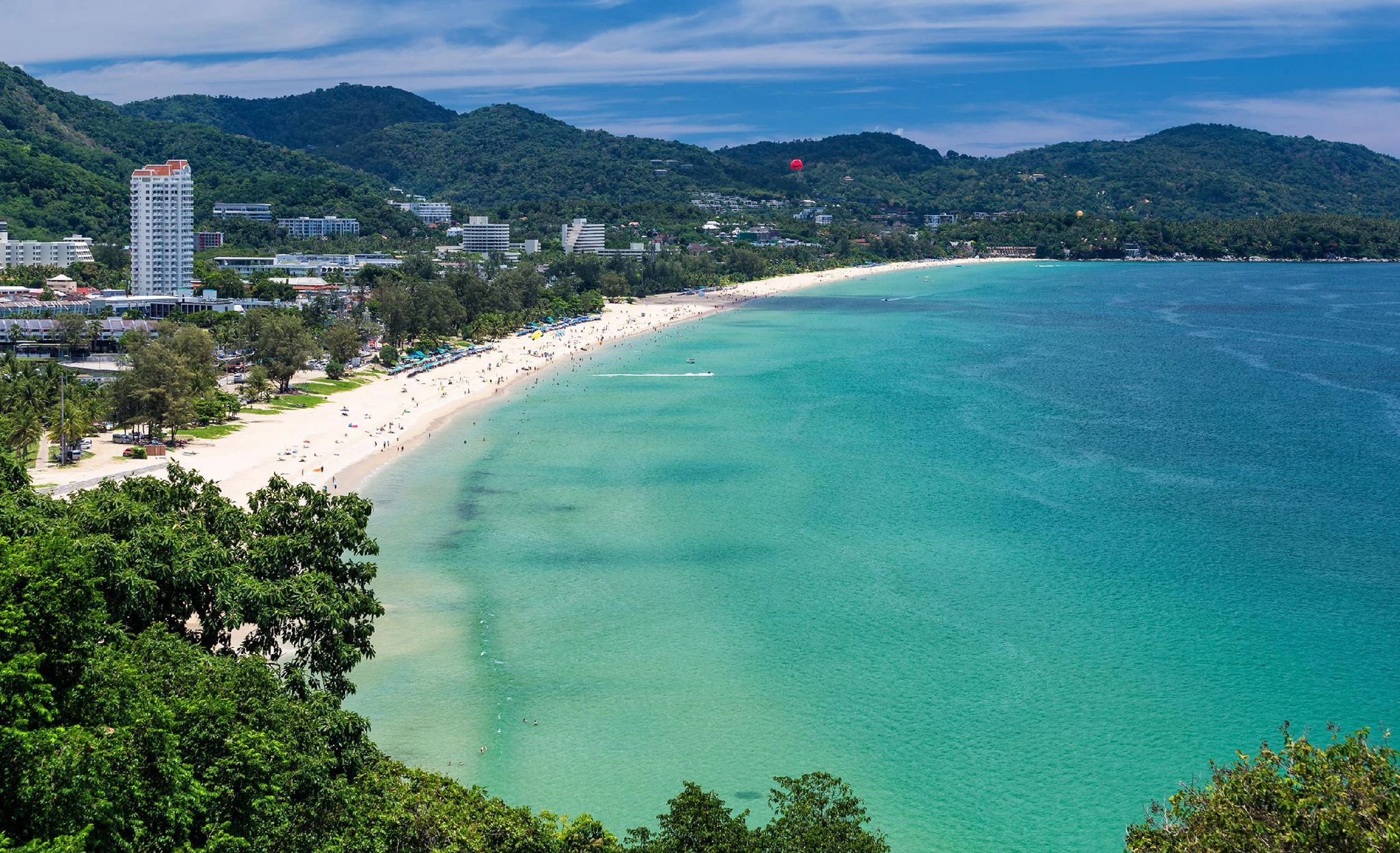 Panoramic view of Karon Bay from above: long sandy shoreline, turquoise sea, and hotels at the foot of green mountains