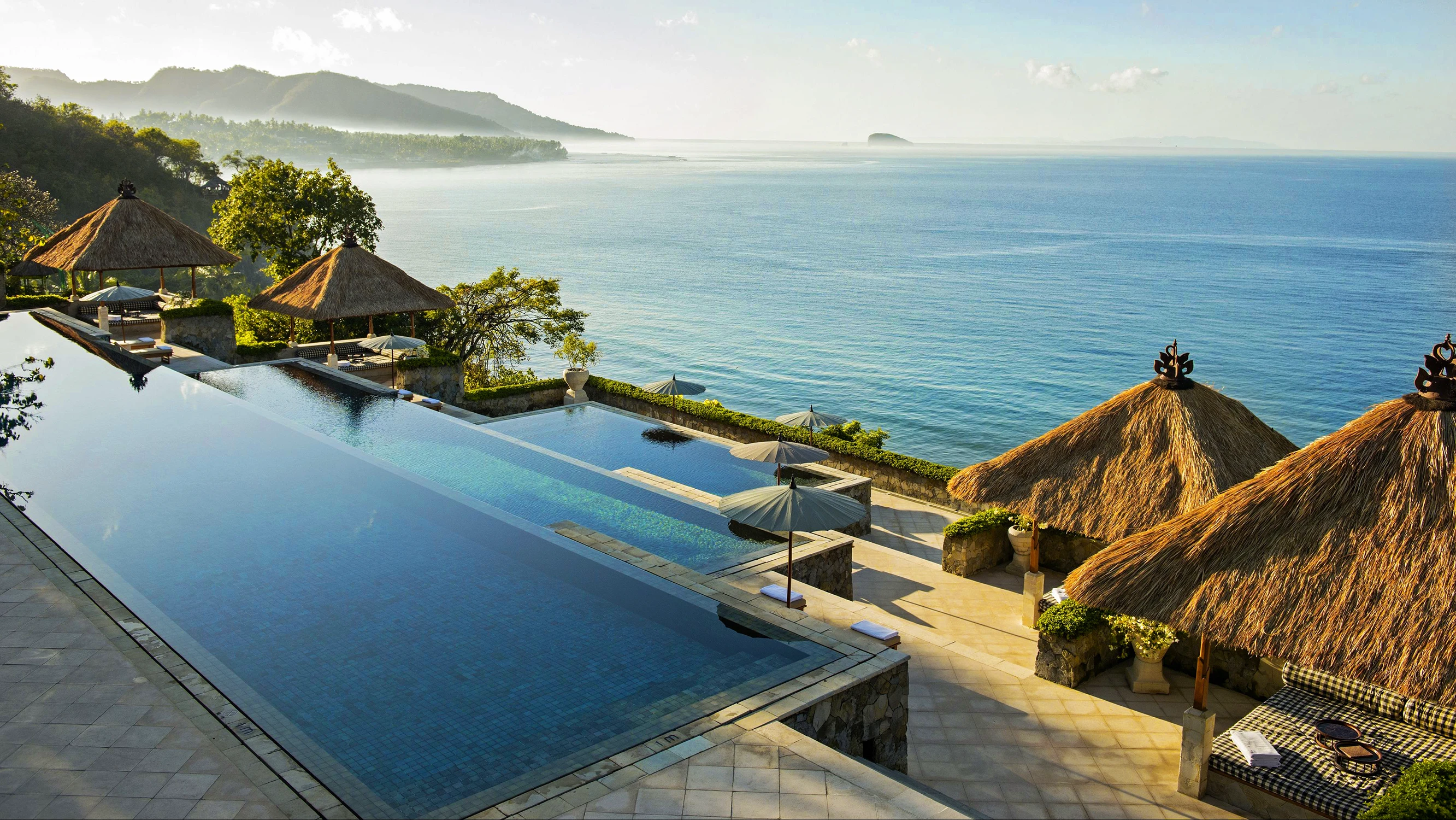 Aerial view of a luxury Bali resort featuring tiered infinity pools, thatched gazebos, and a panoramic ocean view in the morning mist